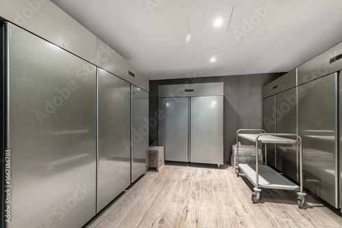Spacious storage area features multiple stainless steel refrigerators lined against the wall, with a utility cart on wheels nearby, illuminated by overhead lights