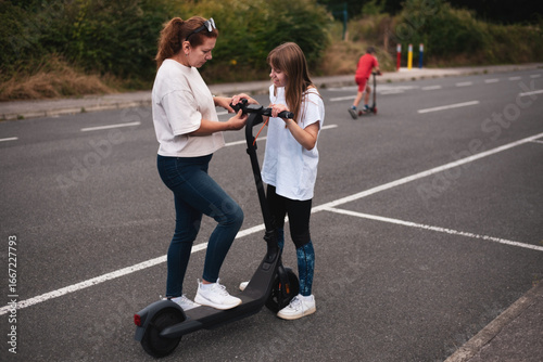 Two individuals engage with an electric scooter on outdoor pavement while one tries it out. Both seem actively immersed in learning and experimenting with the scooter