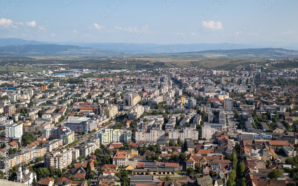Fototapeta premium An elevated, panoramic shot of a city center with dense residential and commercial buildings under a clear sky. In the background, rolling hills and a mountain range can be seen