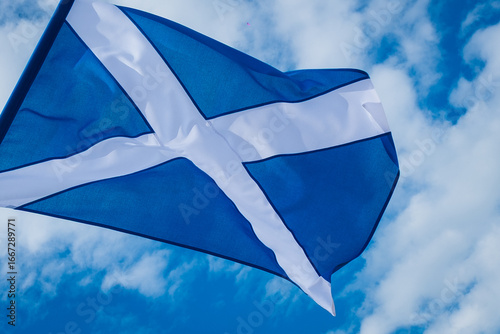 The national flag of Scotland, the Saltire or St. Andrew’s Cross, flying on a flagpole against a blue sky with scattered clouds, symbolizing Scottish heritage, pride, identity. Scottish Flag Waving.