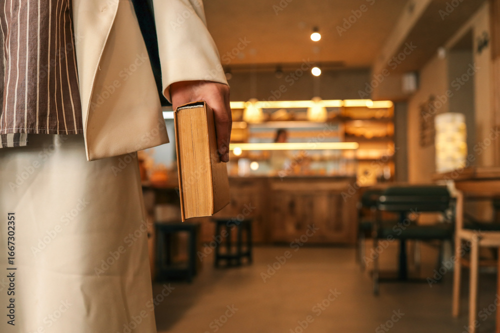 Obraz premium Close-up of woman holding old book in hand, elegant outfit with striped shirt and cream skirt, cozy modern café background with warm lights and wooden furniture
