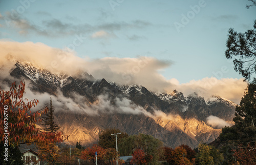 Sun hitting Queenstown snow covered mountains with clouds and a blue sky during autumn time with red and orange leaves