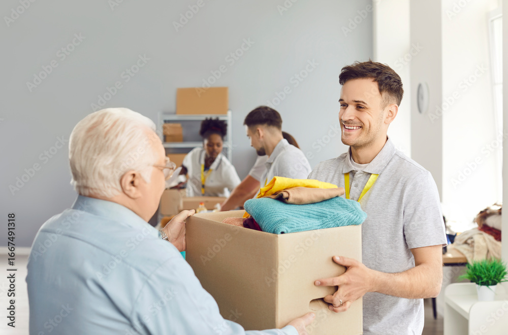 Obraz premium Helpful friendly smiling male volunteer giving senior elderly man box of second hand clothes. Volunteers at humanitarian aid center providing humanitarian help, social support, charity and donations.