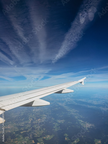 View from an airplane window over the wing, capturing a clear blue sky, a thin contrail, and the vast, blurred patchwork of land below, symbolizing air travel, freedom, and distant destinations.