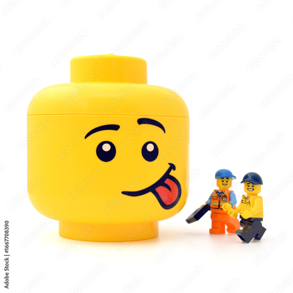 Fototapeta premium Lego minifigures are showing at the large yellow head with funny face is sticking out the tongue isolated on white. Editorial illustrative image of popular plastic toy.