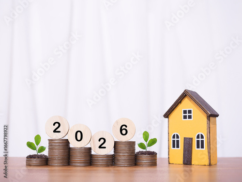 Miniature house, Wooden blocks with number 2026 and plants growing up on stack of coins. The concept of saving money for house, Property investment, House mortgage, Real estate in year 2026.