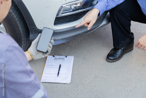 Mechanic taking photo of car damage while customer points at scratch, automotive inspection process for insurance claim or repair documentation, professional service detail in workshop scene