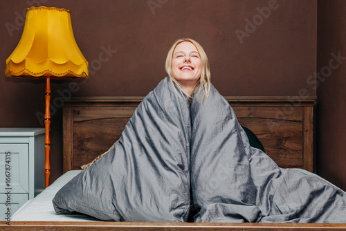 Smiling woman wrapped in gray blanket waking up in cozy bedroom