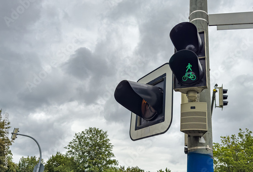 A pedestrian and bicycle signal is visible at an urban intersection under a cloudy sky. The green symbols indicate safe passage for both walkers and cyclists in the area.