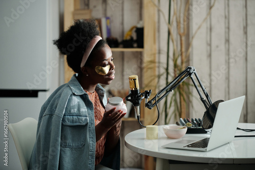 Side view portrait of stylish African American beauty blogger sitting in front of laptop with patches under eyes recording review on beauty product