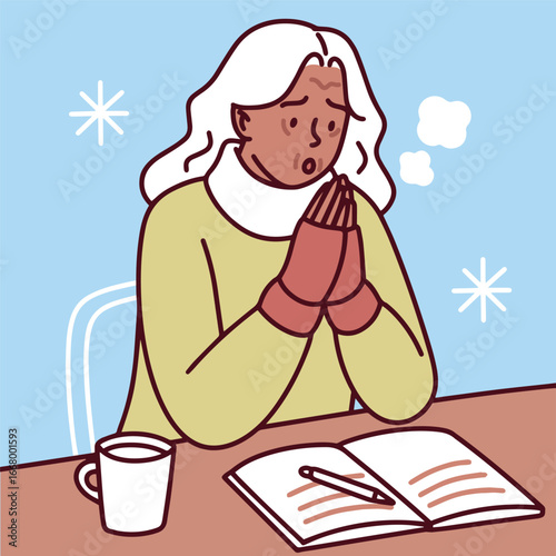Dark-skinned elderly woman with white hair studies at table in cold unheated room holding warm mug while snow falls outside