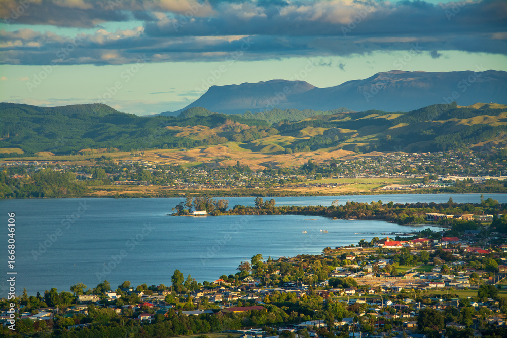 Fototapeta premium A breathtaking view of Rotorua city nestled beside Lake Rotorua, with rolling green hills and distant mountains under a dramatic sky, showcasing the harmony of nature and community