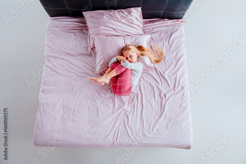 A young girl is curled up in a ball on a bed with pink sheets and pillows. The bright, airy room has natural light illuminating the scene, creating a serene atmosphere.