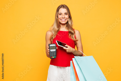 Young woman wears red casual clothes hold shopping package bags bank payment terminal process acquire mobile cell phone isolated on plain orange yellow background. Black Friday sale buy day concept.