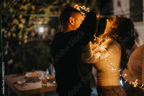 Cuadro en lienzo A couple joyfully entangled in decorative string lights, sharing a tender moment together in the evening outdoors, exuding warmth, happiness, and intimacy