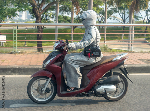 A woman fully covered in hooded clothing rides a red scooter along an urban street.