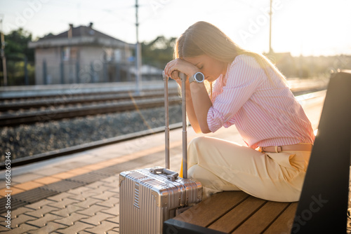 Exhausted businesswoman with luggage sitting on a bench and waiting for delayed transportation at the railway station.	