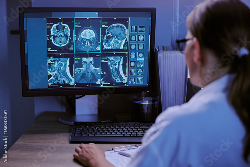 Caucasian middle aged woman analyzing brain and spinal MRI scans on computer monitor in medical office, reviewing detailed radiology images for diagnosis and patient assessment