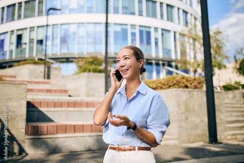 Billede på lærred Asian Business Woman Smiling While Taking a Call Outdoors Near Modern Building