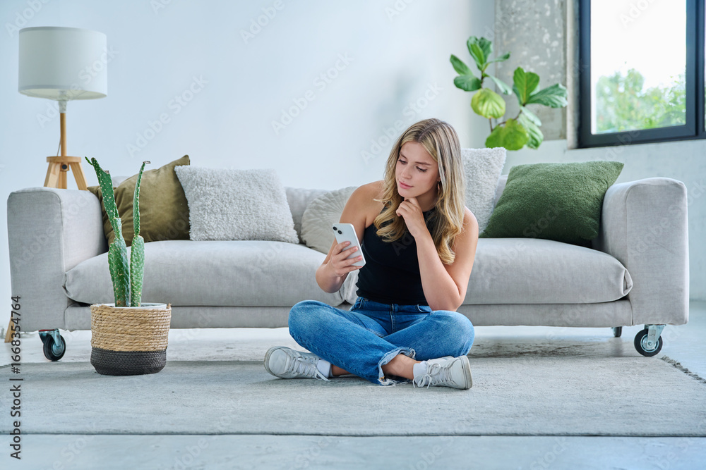 Obraz premium Smiling young woman with smartphone sitting at home near sofa