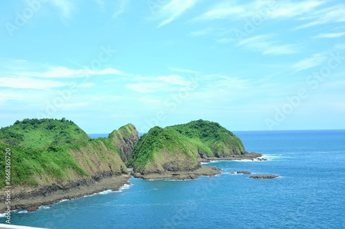 A beautiful coastal landscape at Bedil Island, Banyuwangi, Indonesia. Lush green hills covered in dense vegetation rise dramatically from the clear blue ocean