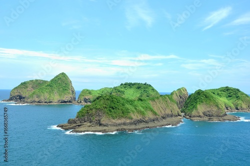 A beautiful coastal landscape at Bedil Island, Banyuwangi, Indonesia. Lush green hills covered in dense vegetation rise dramatically from the clear blue ocean