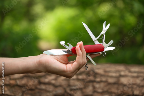 Person holding Swiss army knife in green forest background, vibrant summer tones. Travel and hikink concept