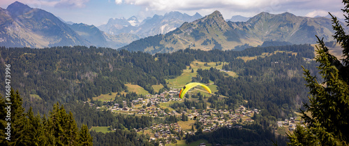 Flying paraglider passing by in stunning French Alps scenery with Mont Blanc massive in the background and Les Gets town in valley below. Anamorphic cinematic panorama.