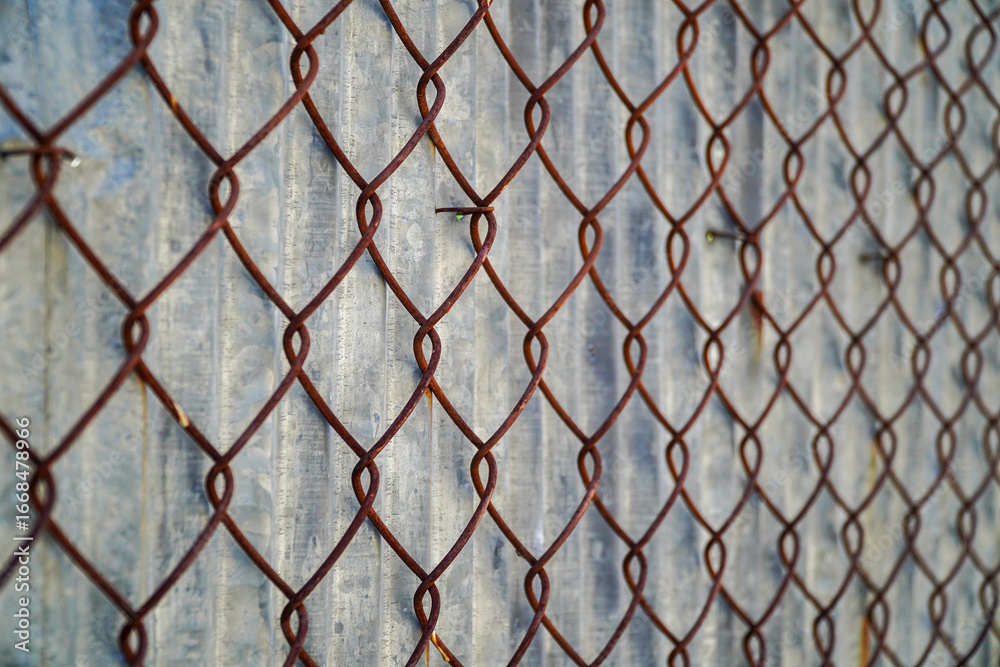 Fototapeta premium A rusty chain-link fence creates a textured and geometric pattern against a corrugated metal background. The play of light and shadow adds depth and visual interest to this abstract composition.