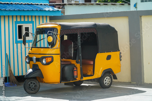 Fotografia A vibrant yellow auto rickshaw contrasts with a striped building, embodying culture and colorful simplicity