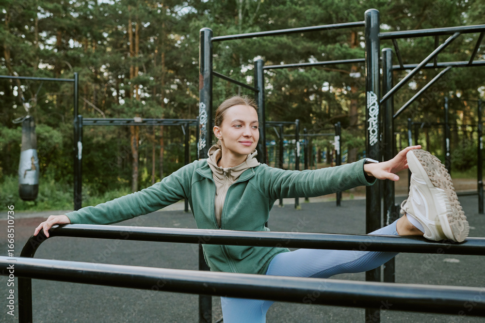Fototapeta premium Caucasian young adult woman stretching leg on outdoor fitness equipment in park, smiling and looking away from camera, surrounded by trees and exercise structures