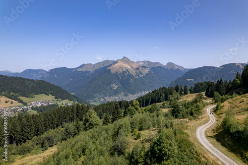 Tableau sur toile Aerial mountainous landscape with meandering path through lush green French Alps valley tourist destination winter sports near Les Gets village with rocky mountain top against a clear blue sky