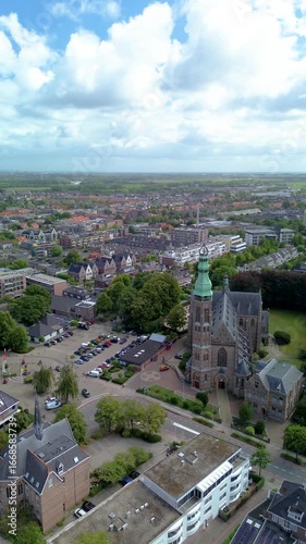 Aerial view of a historic church with a tall brick tower, surrounded by trees, houses, and streets in a small European town. Aerial view of a charming European town with red rooftops, green trees.