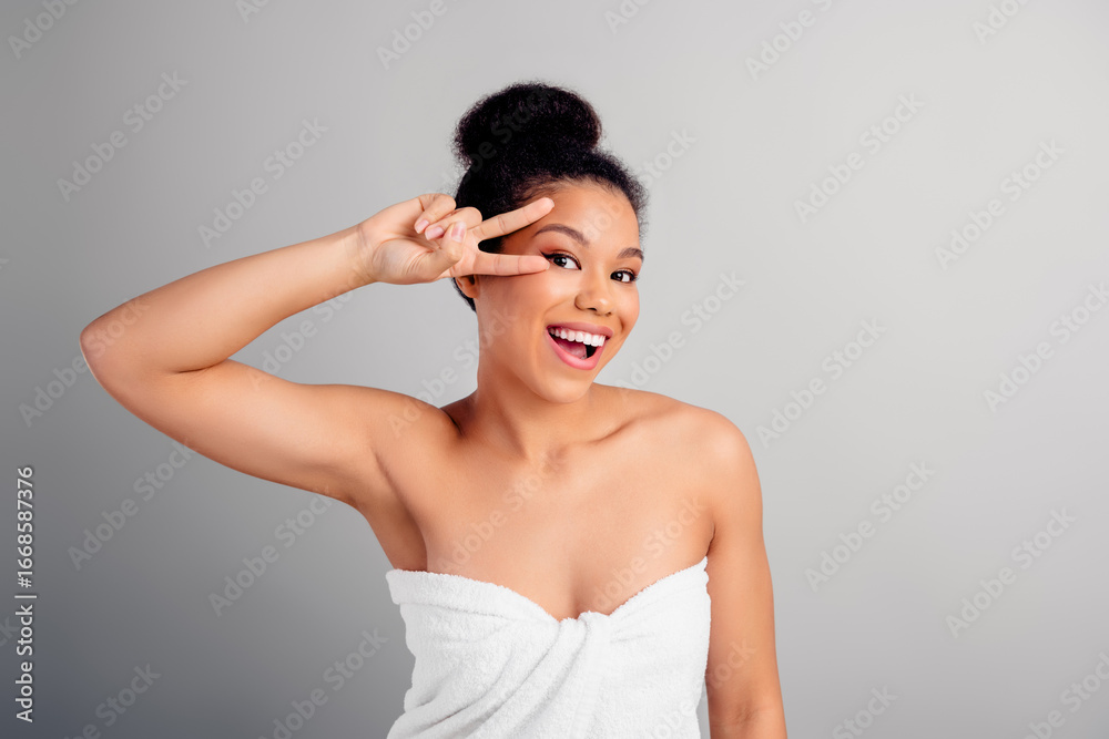 Obraz premium Cheerful young woman with a radiant smile gesturing peace sign, wearing a towel, standing against a white background