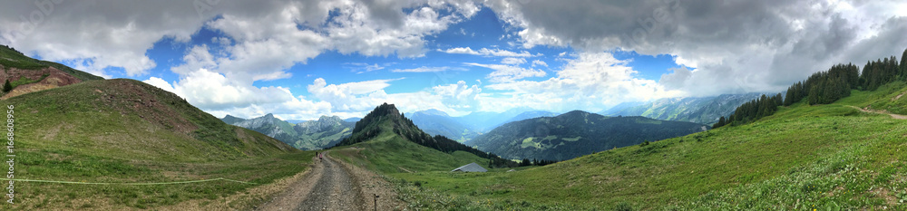 Panoramic mountain view with hiking trail under bright sky and scattered clouds Panoramic mountain view with hiking trail under bright sky and scattered clouds