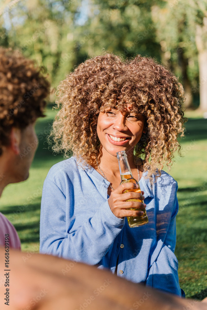 Fototapeta premium Curly-haired woman laughing, gripping beer bottle while enjoying sunny park hangout with close friend, radiating happiness during leisurely outdoor gathering