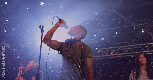 Singer wearing T-shirt singing on club stage under metal truss with headphones, mic and confetti