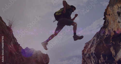 Leaping hiker wearing wide-brimmed hat and backpack crossing rocky canyon gap under sun flare