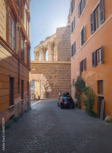 ROME, ITALY - AUGUST 01, 2025 view down a cobblestone roman street, leading to ancient ruins. The path is flanked by historic buildings with shuttered windows. The sun backlights classical columns