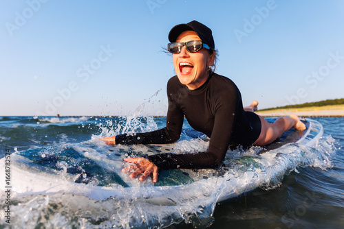 Sticker Smiling woman enjoys surfing on Baltic Sea waves, lying on board with splashing water around