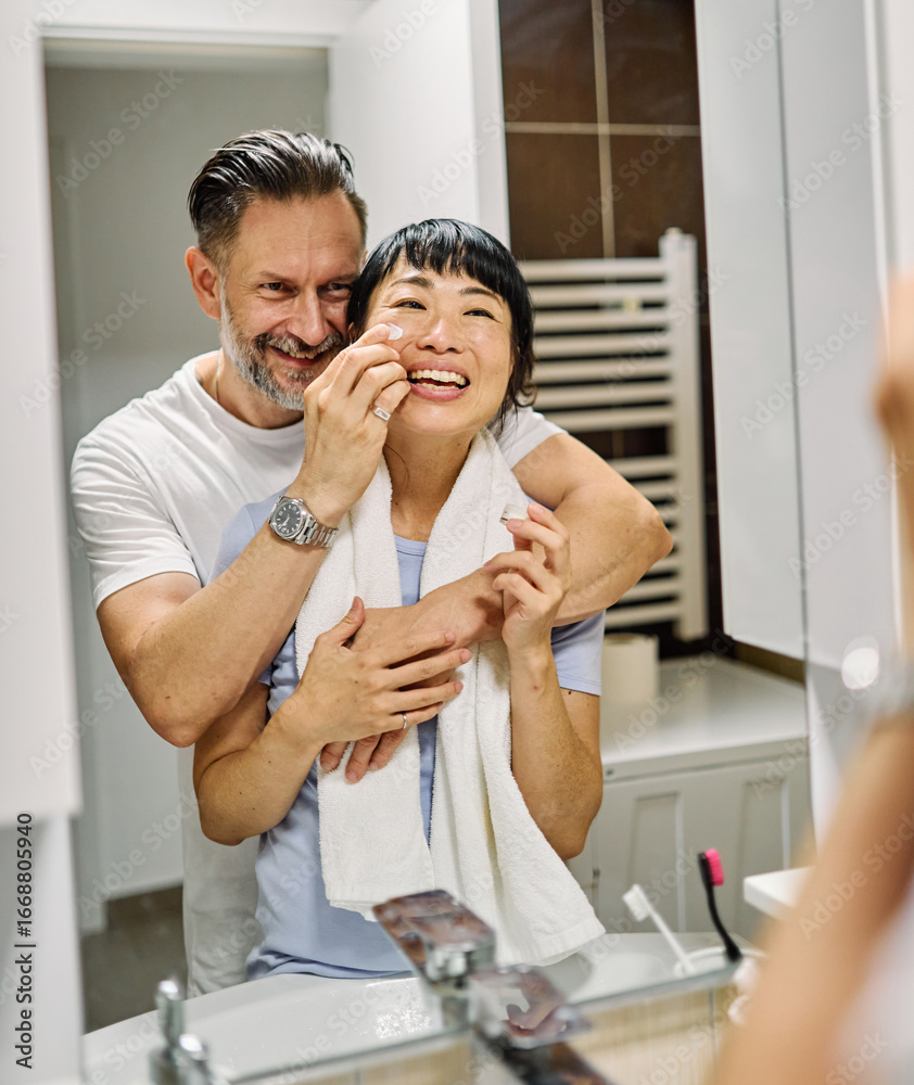 Fototapeta premium Portrait of a young couple applying cream, skincare and cosmetic, in front of mirror in bathroom. Dental hygiene, youth, love and beauty concepts