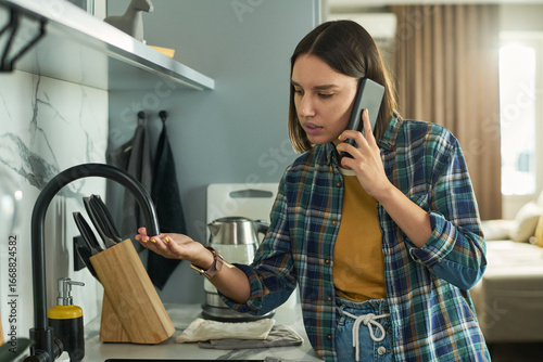 Fototapete Young adult Caucasian woman standing in kitchen talking on smartphone while chec