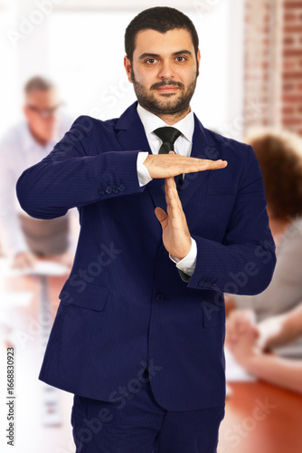 Confident businessman in a suit making a timeout gesture during a group office meeting, symbolizing the need for a break or pause. In the background, colleagues are engaged in discussion at the table.