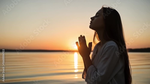 Young Woman in White Shirt Praying Silhouetted Against a Golden Sunset Sky Over Calm Waters During Dusk Peaceful Landscape
