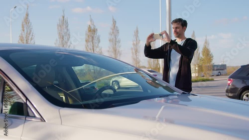 Man taking smartphone photo of car in parking lot, showcasing carsharing experience with modern vehicles and clear blue sky in the background, capturing urban lifestyle