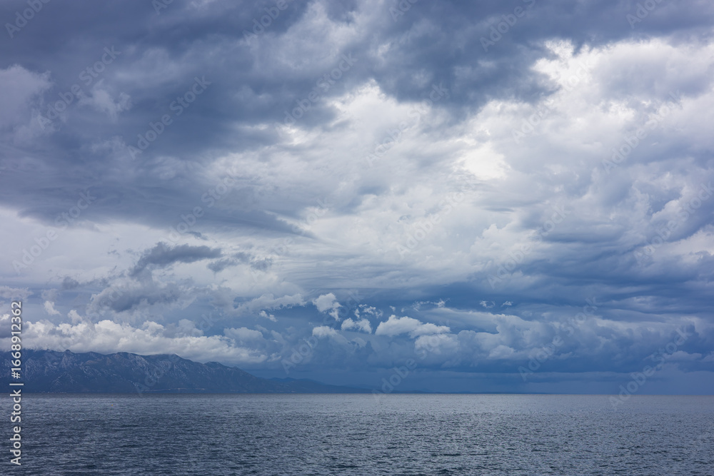 Obraz premium Stormy sky over mountains and sea. Dark dramatic clouds hang over the water surface, creating an atmosphere of tension and grandeur of nature. Podaca village, Makarska Riviera, South Dalmatia, Croatia