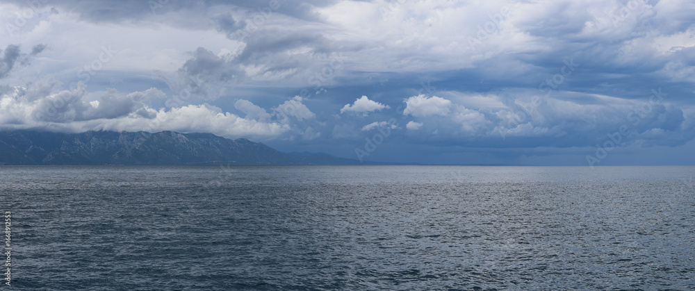 Fototapeta premium Stormy sky over mountains and sea. Dark dramatic clouds hang over the water surface, creating an atmosphere of tension and grandeur of nature. Podaca village, Makarska Riviera, South Dalmatia, Croatia