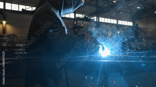 Welder in protective gear working on metal structure in a factory, creating bright sparks and intense light. Industrial manufacturing process in action