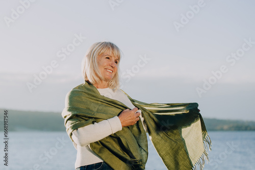 Fotografía Smiling senior woman holding green shawl by the lake enjoying retirement