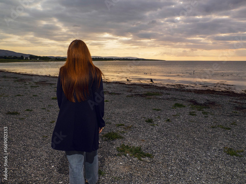 Fotografie Red hair girl in black long coat is walking on a stone beach towards ocean at sunset wit dark and moody sky